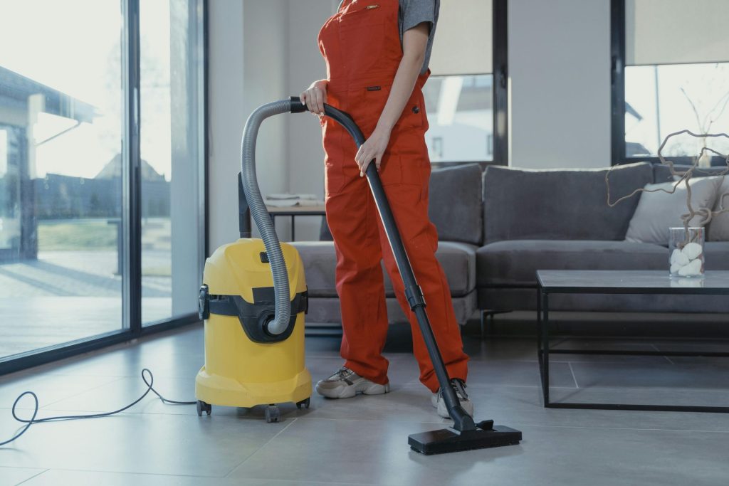 Ein professioneller Reinigungskraft in rotem Overall saugt mit einem gelben Staubsauger ein modernes Wohnzimmer.